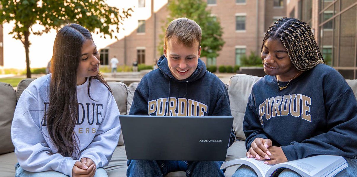 three students sit on a campus bench using a laptop.