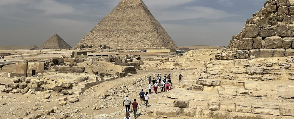 A group of tourists walks along a sandy path toward the Great Pyramids of Giza under a hazy sky, surrounded by ancient stone ruins.